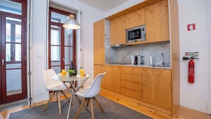 Een kleine keuken met houten kastjes, een magnetron, en een eethoekje met twee stoelen en een tafel.