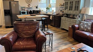 A modern kitchen with a refrigerator, microwave, and coffee machine. There is a dining area with a wooden table and chairs. The kitchen has a window and a cabinet with decorative items.
