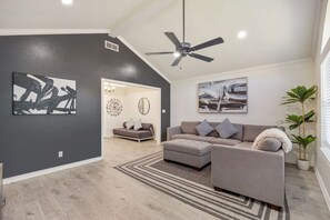 Un salon moderne avec un canapé gris, un fauteuil gris et une table basse. Les murs sont ornés de tableaux abstraits, et on y trouve également un ventilateur de plafond et une plante en pot.