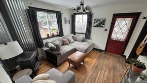 Un salon chaleureux avec un canapé, un pouf et une cheminée. On y trouve des fenêtres avec des rideaux, une porte rouge et une guitare appuyée contre le mur.