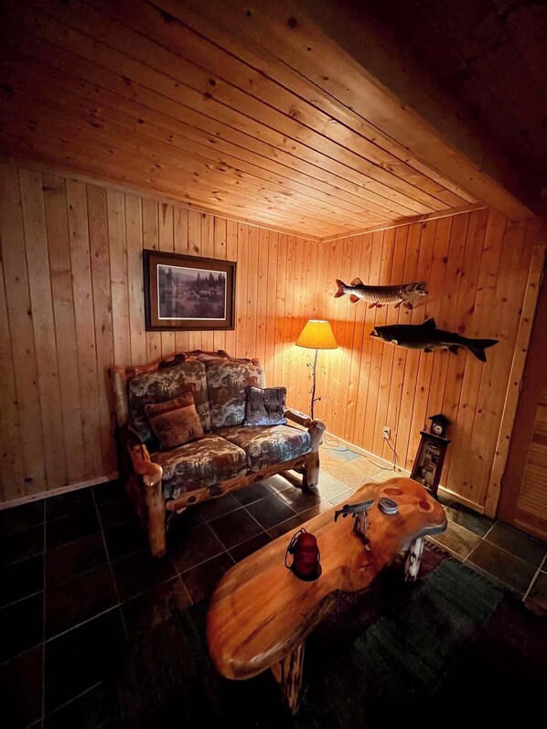 En mysig stuga med träpanel på väggarna, en inramad tavla på väggen, en soffa, en lampa och uppstoppade fiskar som hänger på väggen.
