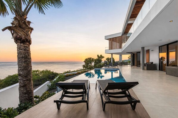 A modern outdoor pool area with a view of the ocean at sunset, lounge chairs, and a contemporary building structure.