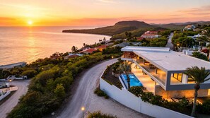 A coastal property with a swimming pool, a road leading to the property, and a sunset view. - Luxury 12-Guest Villa: Pool, Oceanview & Beach (Bandabou)