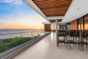 A modern balcony with a view of the ocean, featuring a wooden ceiling, glass railings, and a seating area with tables and chairs.