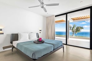 A modern hotel room with a bed, a ceiling fan, a nightstand, a lamp, and a view of the ocean. - Luxury 12-Guest Villa: Pool, Oceanview & Beach (Bandabou)