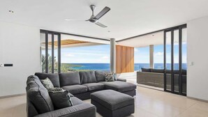 Ein modernes Wohnzimmer mit einer Eckcouch, einem kleinen Hocker und einem Deckenventilator. Das Zimmer verfügt über große Schiebeglastüren, die auf einen Balkon mit Meerblick führen.