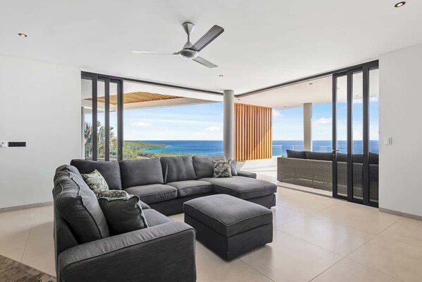 A modern living room with a sectional sofa, a small ottoman, and a ceiling fan. The room has large sliding glass doors leading to a balcony with a view of the ocean.