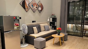 A modern living room with a sofa, a small round table with flowers, a standing fan, and a refrigerator. - Sandy Homestay Citadines Marina Ha Long is a great choice for accommodation (Ha Long)