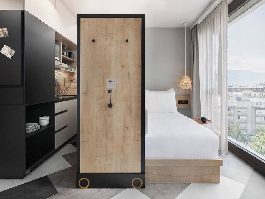 A modern hotel room with a large bed, a wardrobe, a desk, and a view of the city.