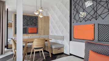 Una habitación moderna con una mesa de comedor de madera, dos sillas, un banco y un sofá. Hay un estante fijado a la pared con un diseño geométrico y un bolso pequeño naranja.