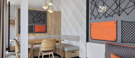 A modern room with a wooden dining table, two chairs, a bench, and a sofa. There is a wall-mounted shelf with a geometric pattern and a small orange bag.