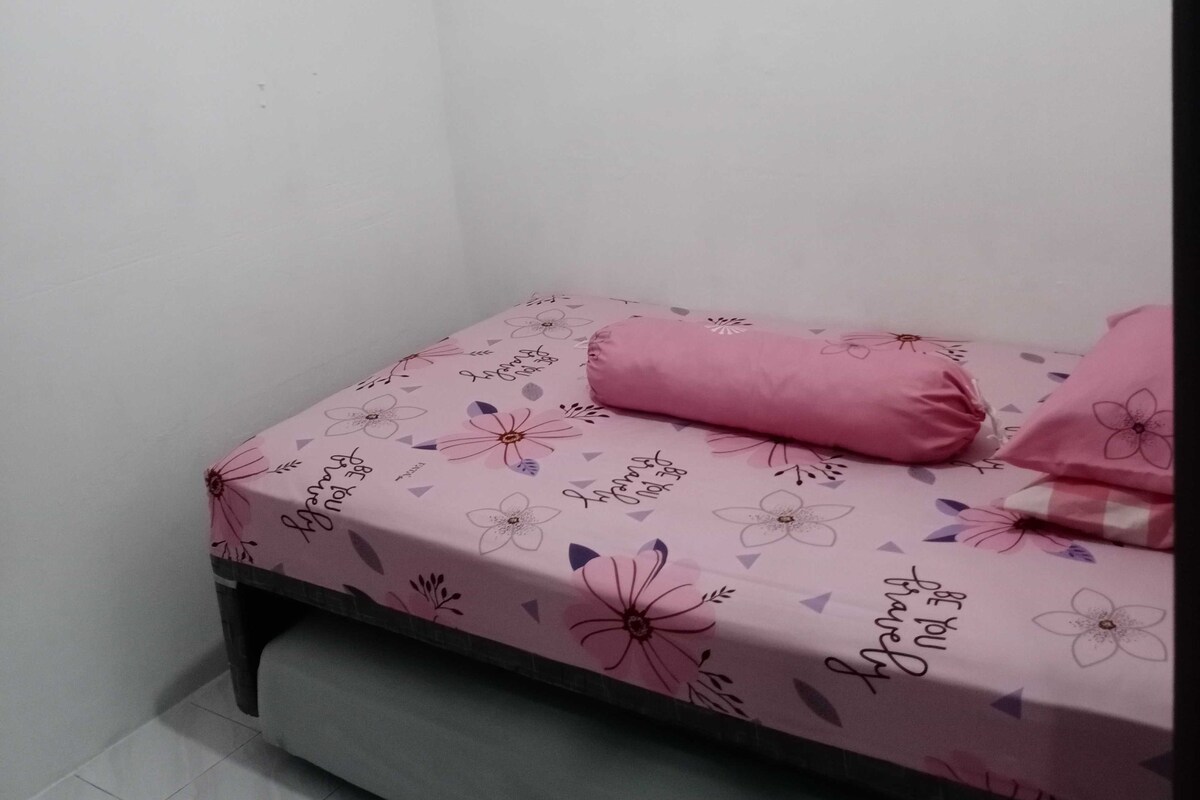 Une petite chambre avec un lit simple, une couvre-lit à motifs floraux roses et des coussins assortis.