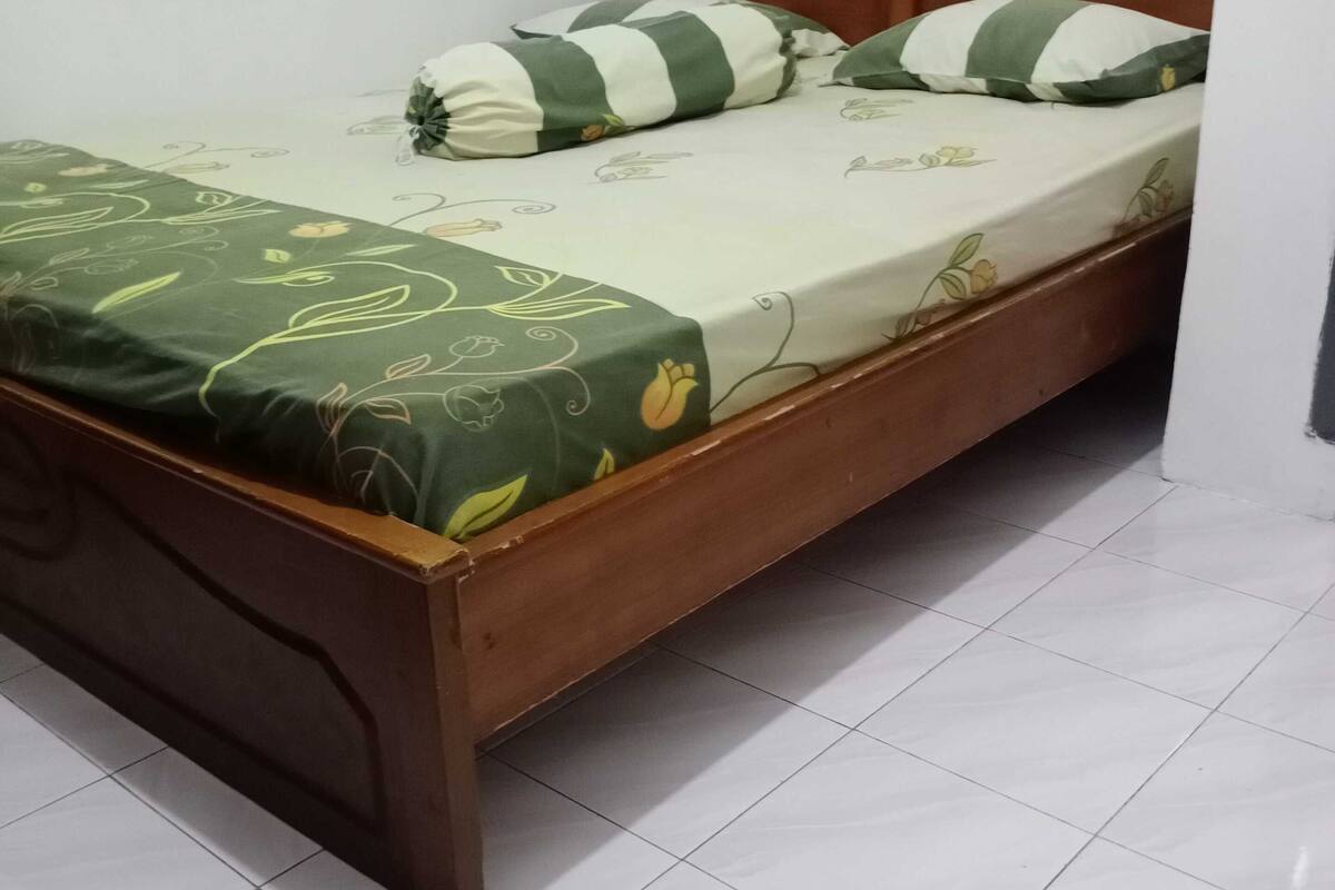 Un lit simple avec un cadre en bois, une couvre-lit à motifs floraux verts et deux coussins à rayures.