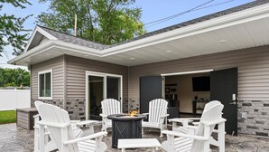 En uteplats med Adirondack-stolar och ett runt bord, ett golv belagt med stengolv och ett hus med en fasad som har en accentvägg av sten och en skjutdörr i glas.