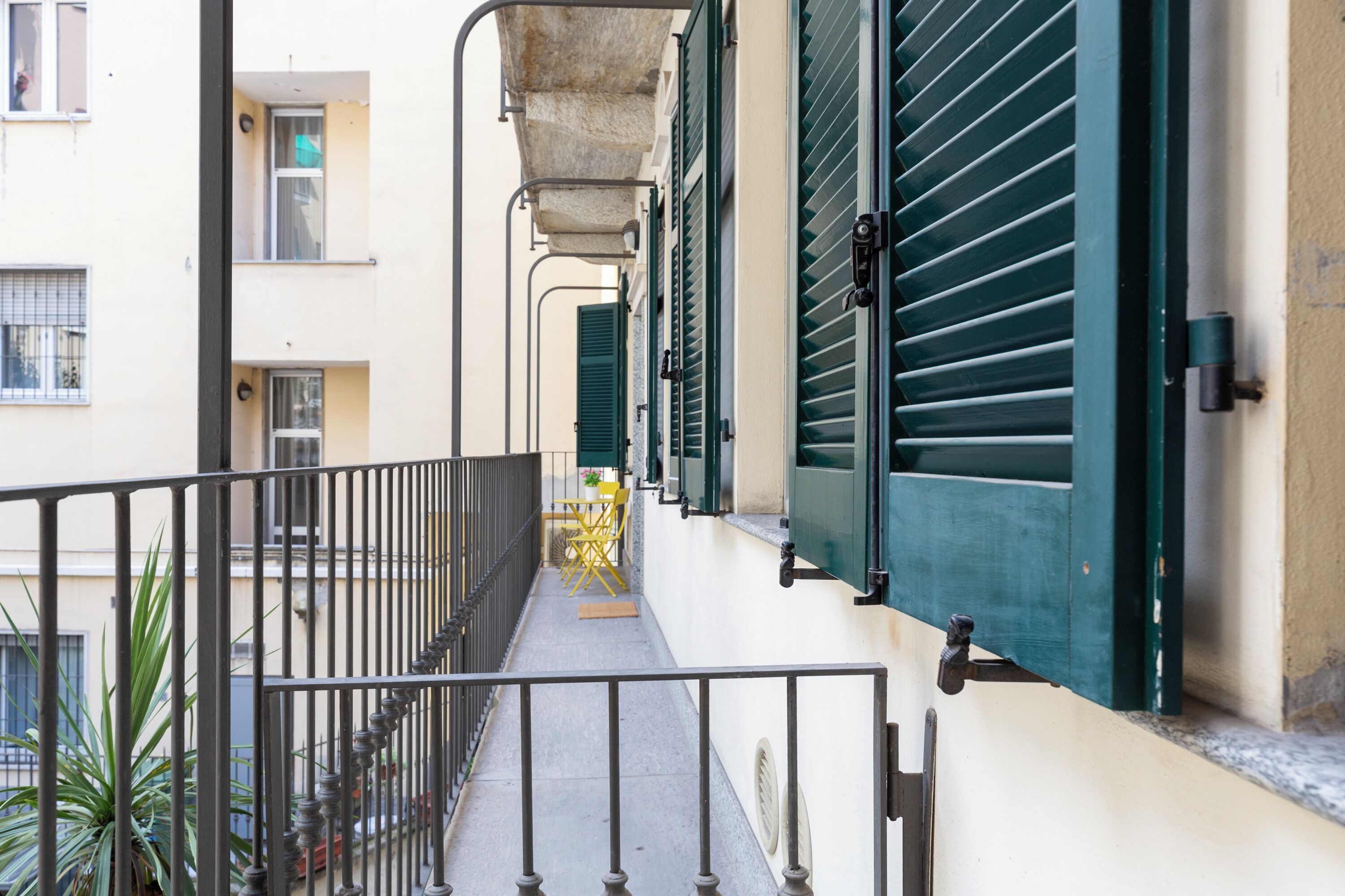 A narrow balcony with a metal railing, green shutters, and a small table with chairs.