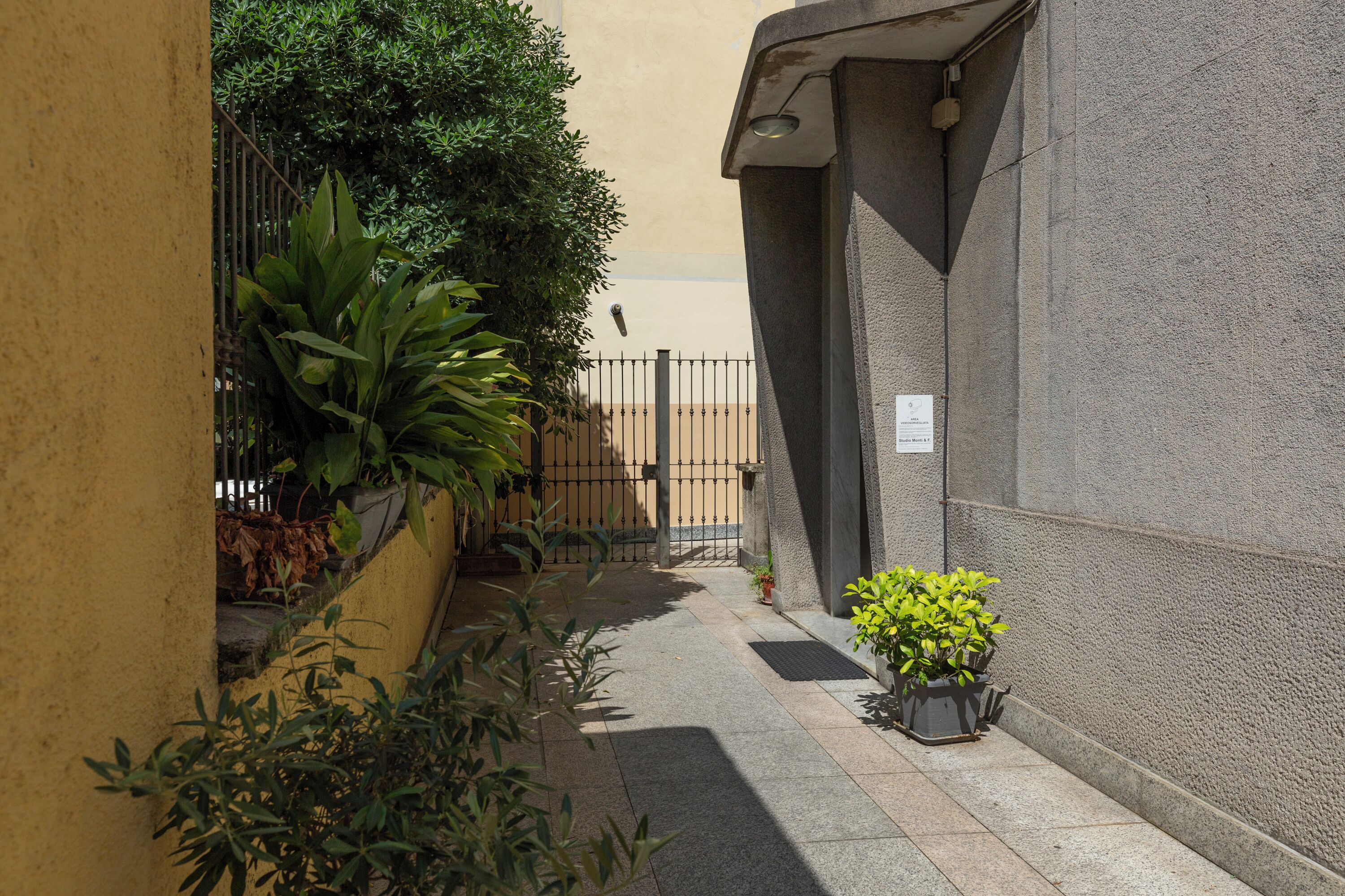 A narrow alleyway with a yellow wall on one side and a grey wall on the other, featuring potted plants and a metal gate.