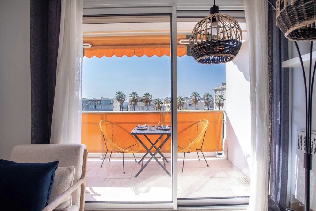 Ein Balkon mit Tisch und Stühlen, einer Hängelampe aus Korbmöbeln und Blick auf Palmen und Gebäude.