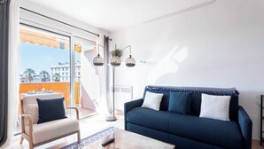 A living room with a sofa, a coffee table, and a chair. There is a balcony with a view of buildings and palm trees. - Cocoon with garden near the sea (Cannes)