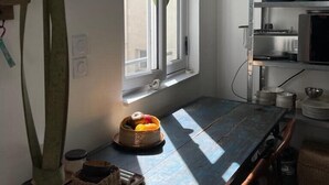 Eine Küche mit Fenster, einem Tisch mit Obstkorb und einem Regal mit Geschirr.