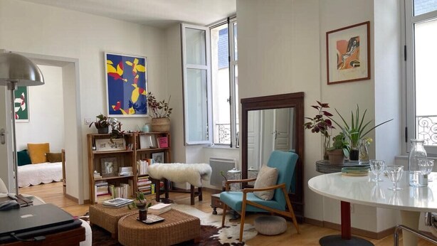 Ein gemütliches Wohnzimmer mit einer Couch, einem Sessel, einem Couchtisch und einem Bücherregal. Ein Fenster bietet Blick ins Freie.