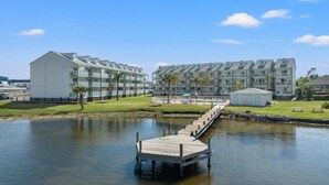Un hôtel situé au bord de l’eau, avec un quai menant à un espace piscine, le tout entouré d’un paysage verdoyant et surplombé d’un ciel bleu azur.