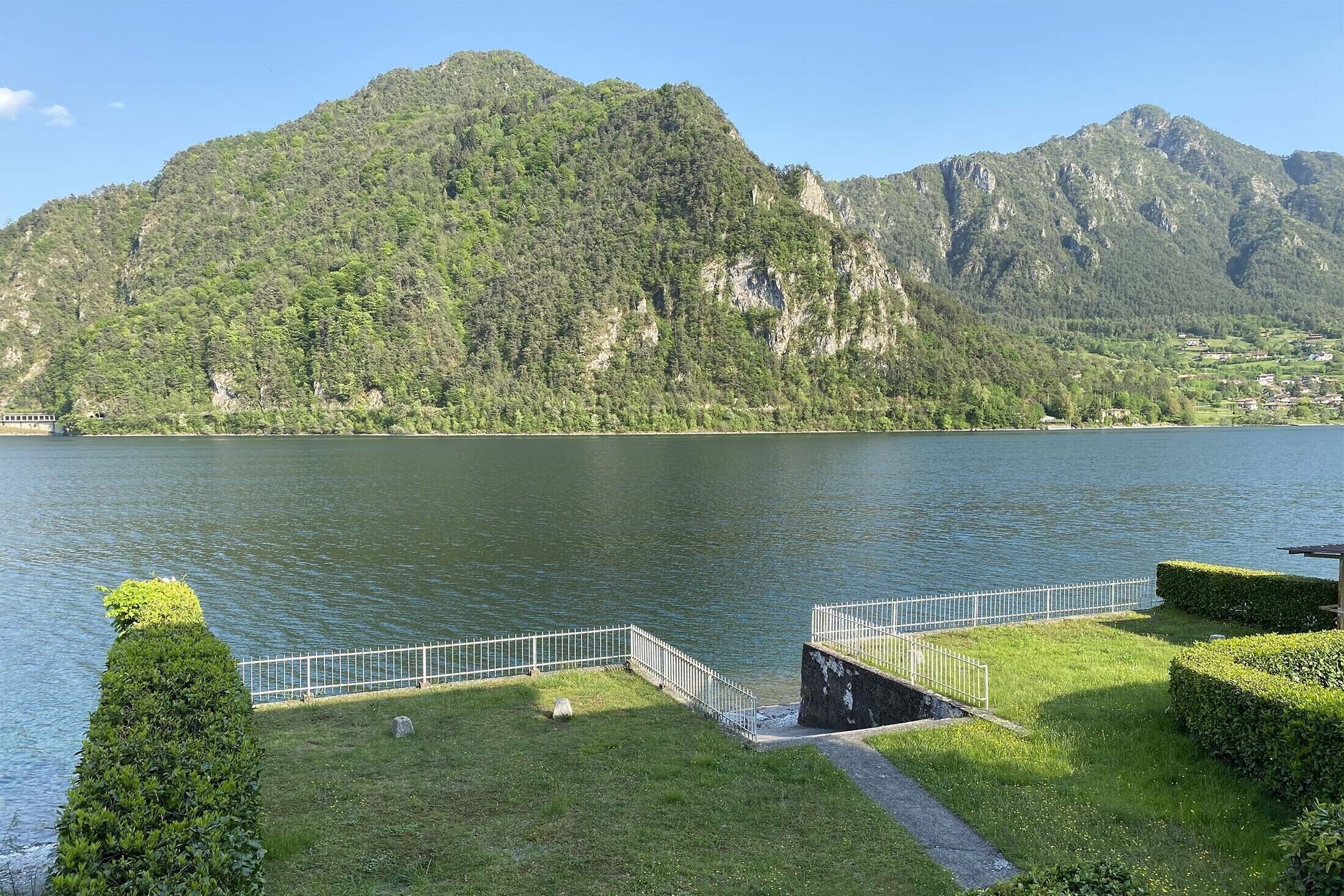 Seeblick mit Bergelandschaft, einer grünen Wiese und einem Metallgeländer am Ufer.