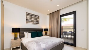 A modern bedroom with a large bed, two bedside lamps, a nightstand, a ceiling light, a view of the outdoors through a sliding door, and a piece of abstract art on the wall.