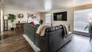 A living room with a dark leather sofa, a flat-screen TV mounted on the wall, and a large window with blinds. - Hoff Gardens 12 - 2BR 2BA - Downtown Lubbock (Lubbock)