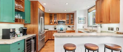 A modern kitchen with wooden cabinets, a built-in oven, a coffee machine, and a bar stool area.