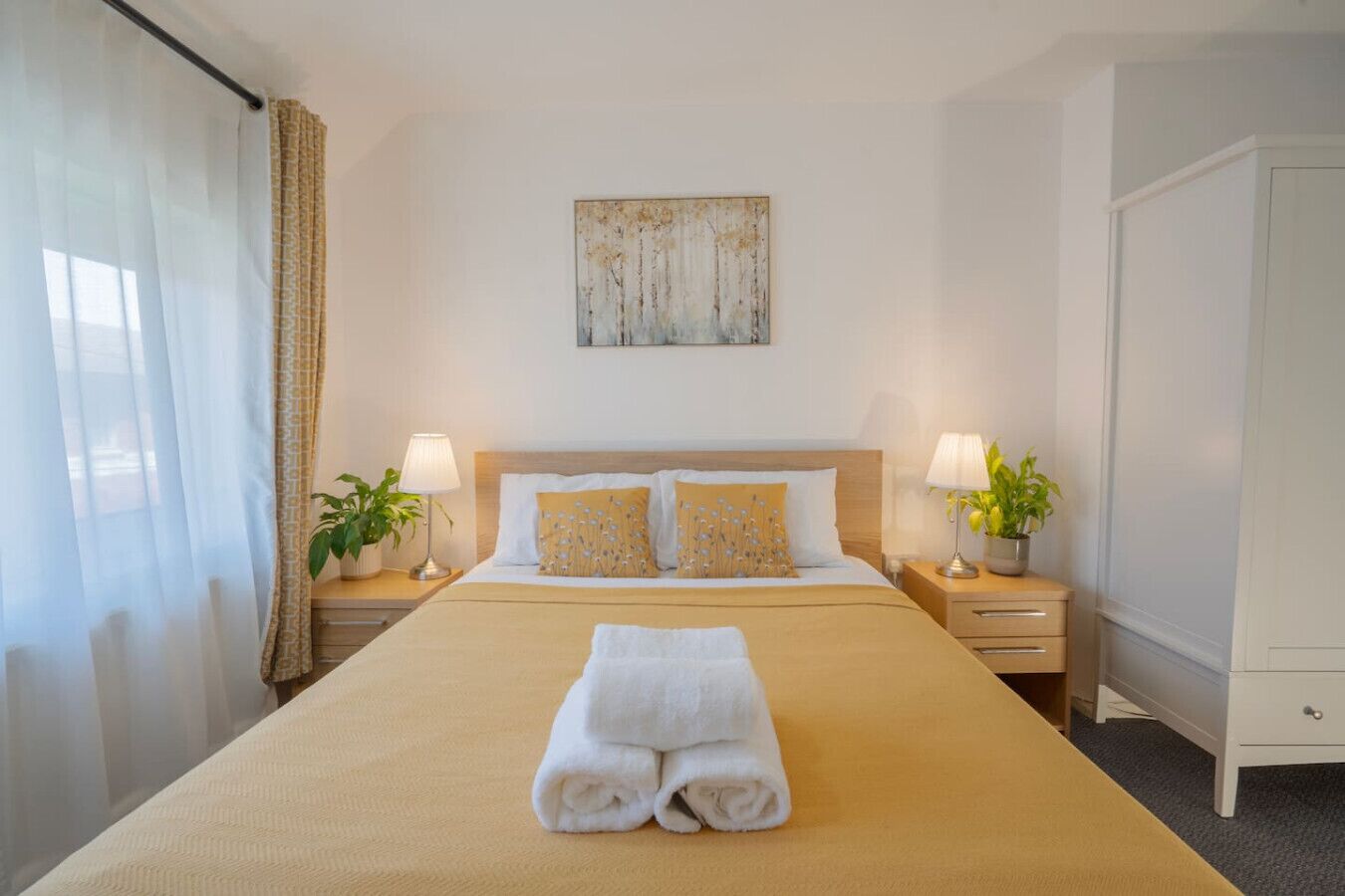 A hotel room with a large bed, two bedside tables with lamps and plants, a wooden headboard, and a painting on the wall.