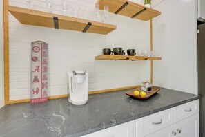Una cocina con cafetera, repisas con vasos y tazas, y un letrero decorativo.