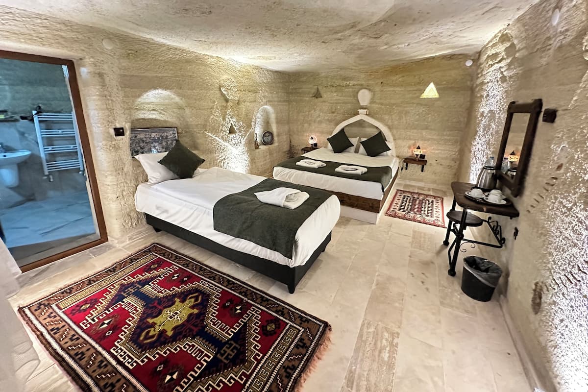 A room with two beds, a small table with cups and a kettle, a rug, and a view of the outside.