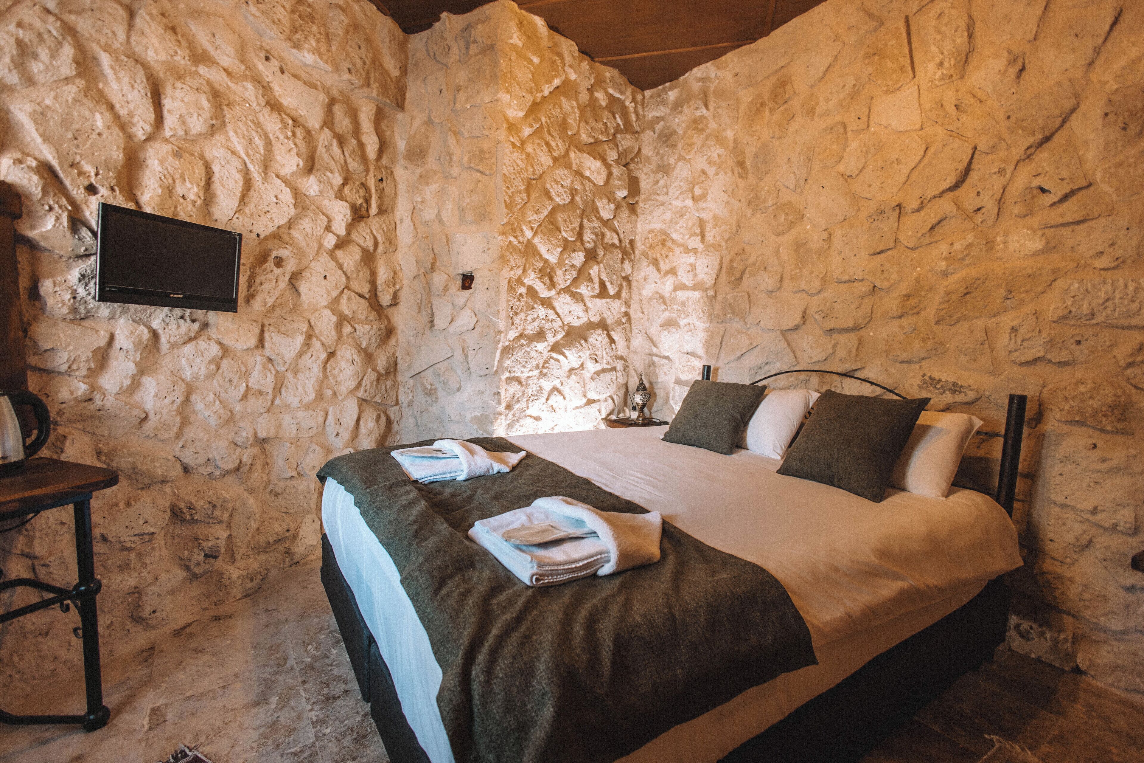 A stone-walled bedroom with a bed, pillows, a flat-screen TV, and a table with a kettle.