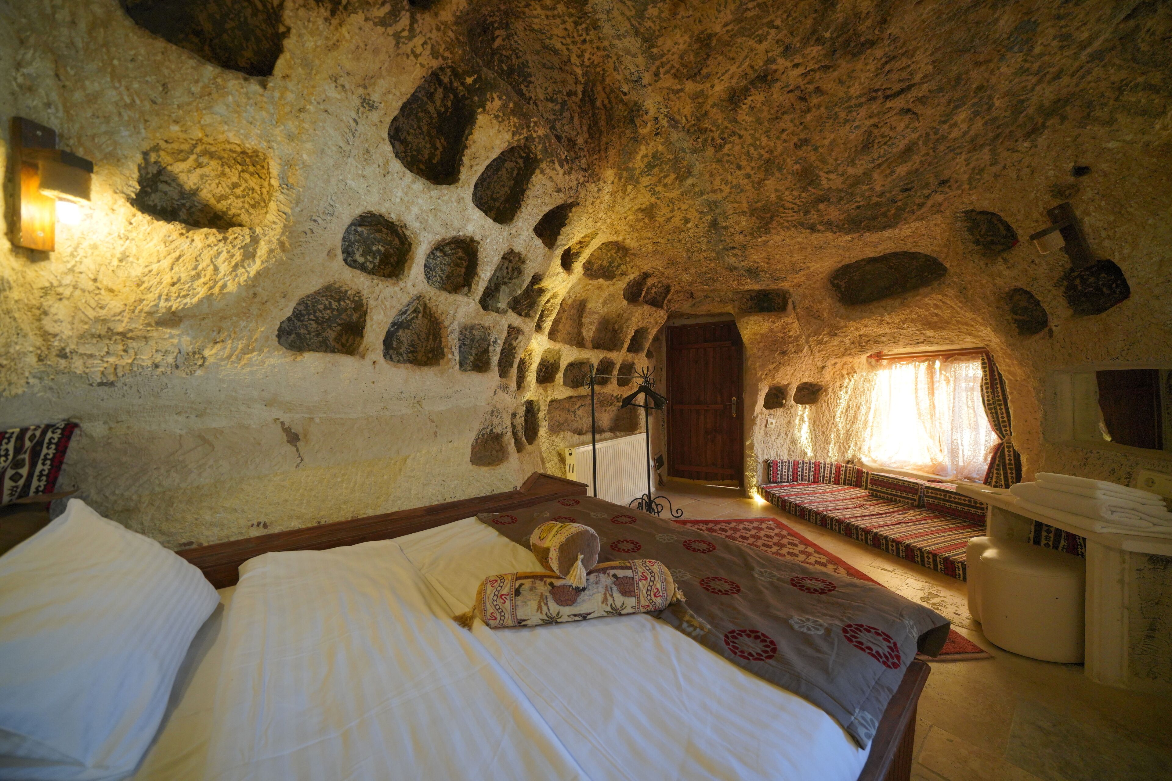 Una habitación rústica de piedra con una cama, una zona de lectura con libros y una puerta de madera.