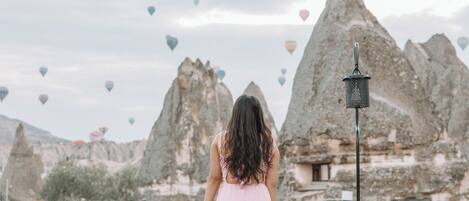 A person in a pink dress standing on a balcony with a view of rocky mountains and hot air balloons.