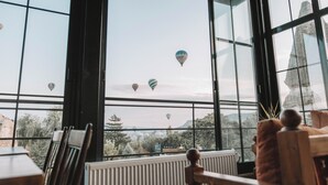 A view through large windows of hot air balloons in the distance, with a rustic interior featuring wooden furniture and a stone sculpture. - TURAN Cappadocia Cave (Nevsehir)