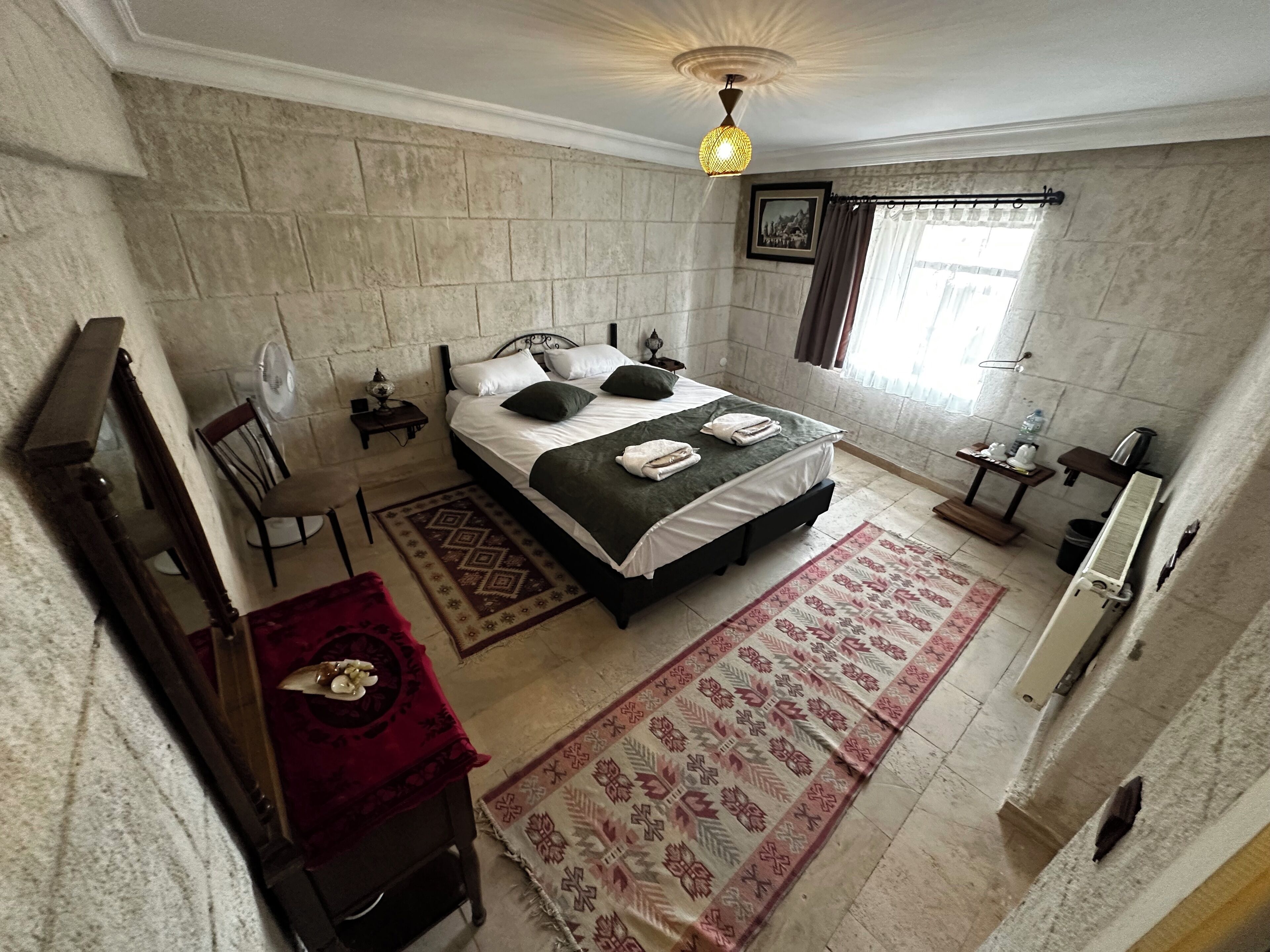 Un dormitorio con cama, sillón, mesita de noche, lámpara, radiador y ventana con cortinas.