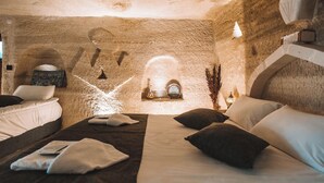 A room with a stone wall, a bed with white and dark bedding, a small table with a lamp, and a decorative wall piece. - TURAN Cappadocia Cave (Nevsehir)