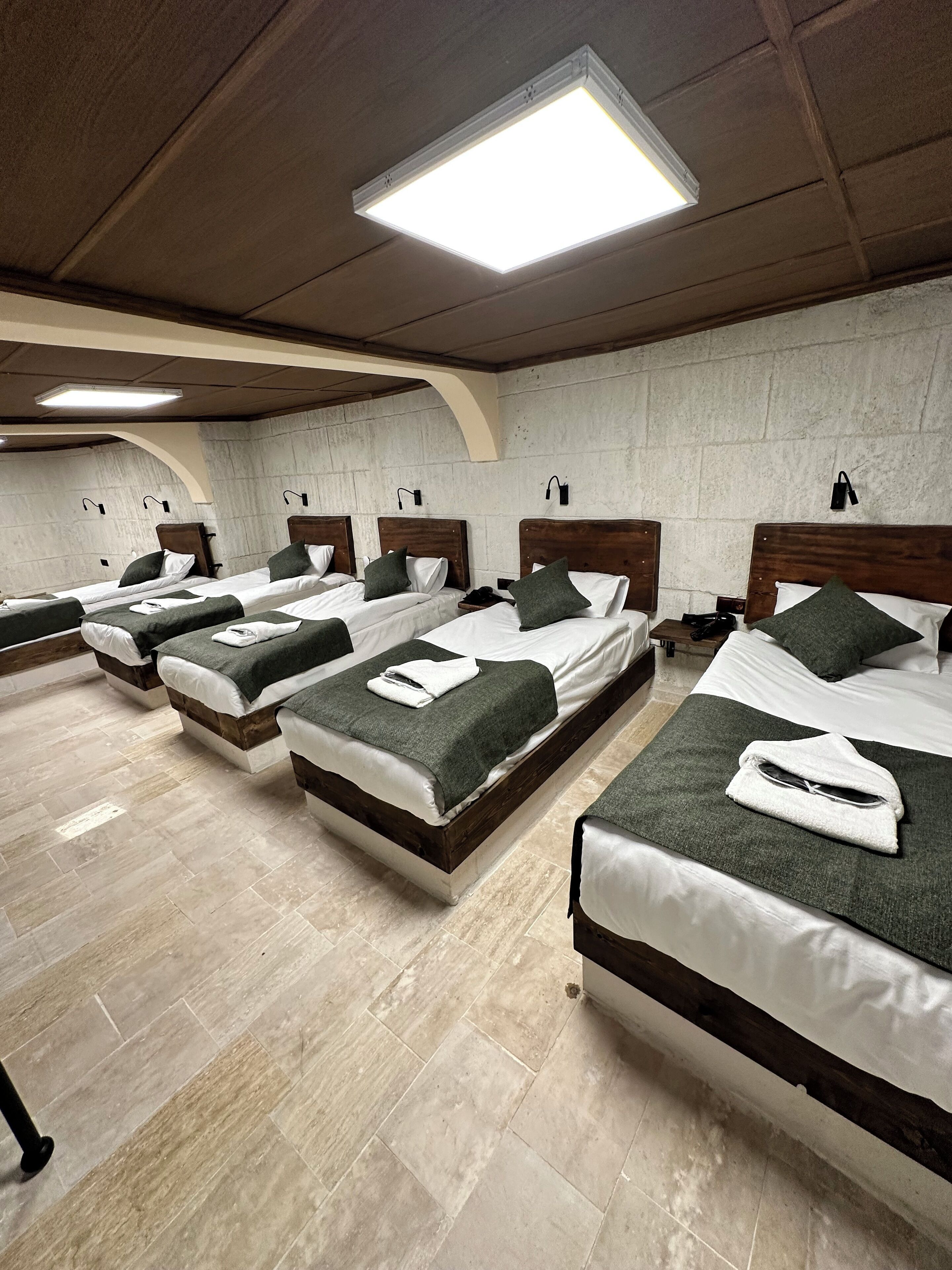 Une chambre d’hôtel avec quatre lits alignés, chacun doté d’une tête de lit en bois, de draps blancs et de coussins décoratifs verts. Un grand luminaire est fixé au plafond, et les murs sont en béton texturé.