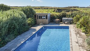 Une piscine rectangulaire à l’eau d’un bleu limpide, entourée d’une terrasse en pierre. On y trouve une petite construction en bois avec une porte et des fenêtres, ainsi qu’une table et des chaises à proximité.