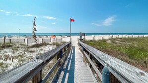 Una pasarela de madera que lleva a una playa con una bandera roja, dunas arenosas y un cielo azul despejado.