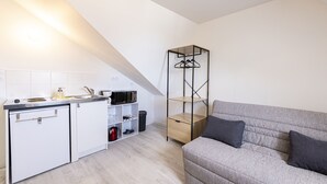 A compact kitchenette with a microwave, a small fridge, and a sink. There's a grey sofa with cushions and a tall, narrow shelving unit against the wall. - L'écrivain - Wifi - Saint-Serge (Angers)
