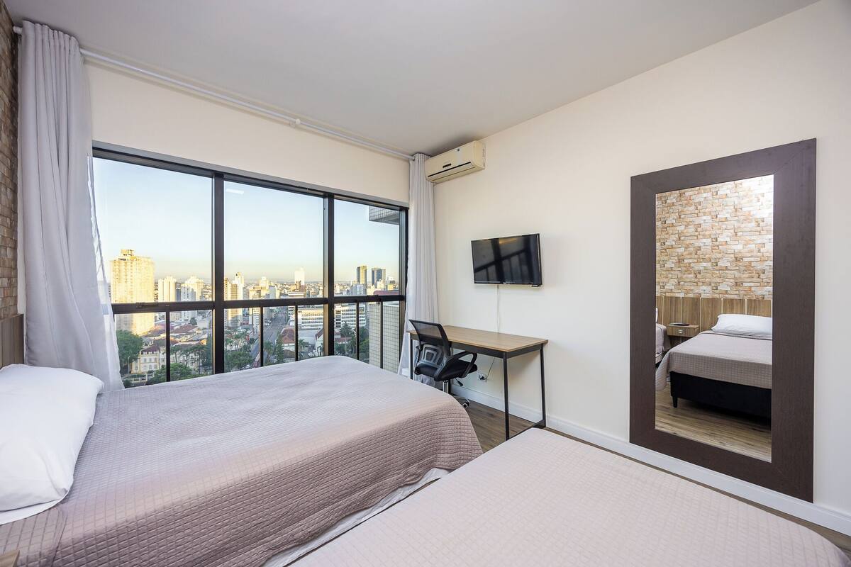A hotel room with two beds, a desk, a TV, and a large window offering a city view.
