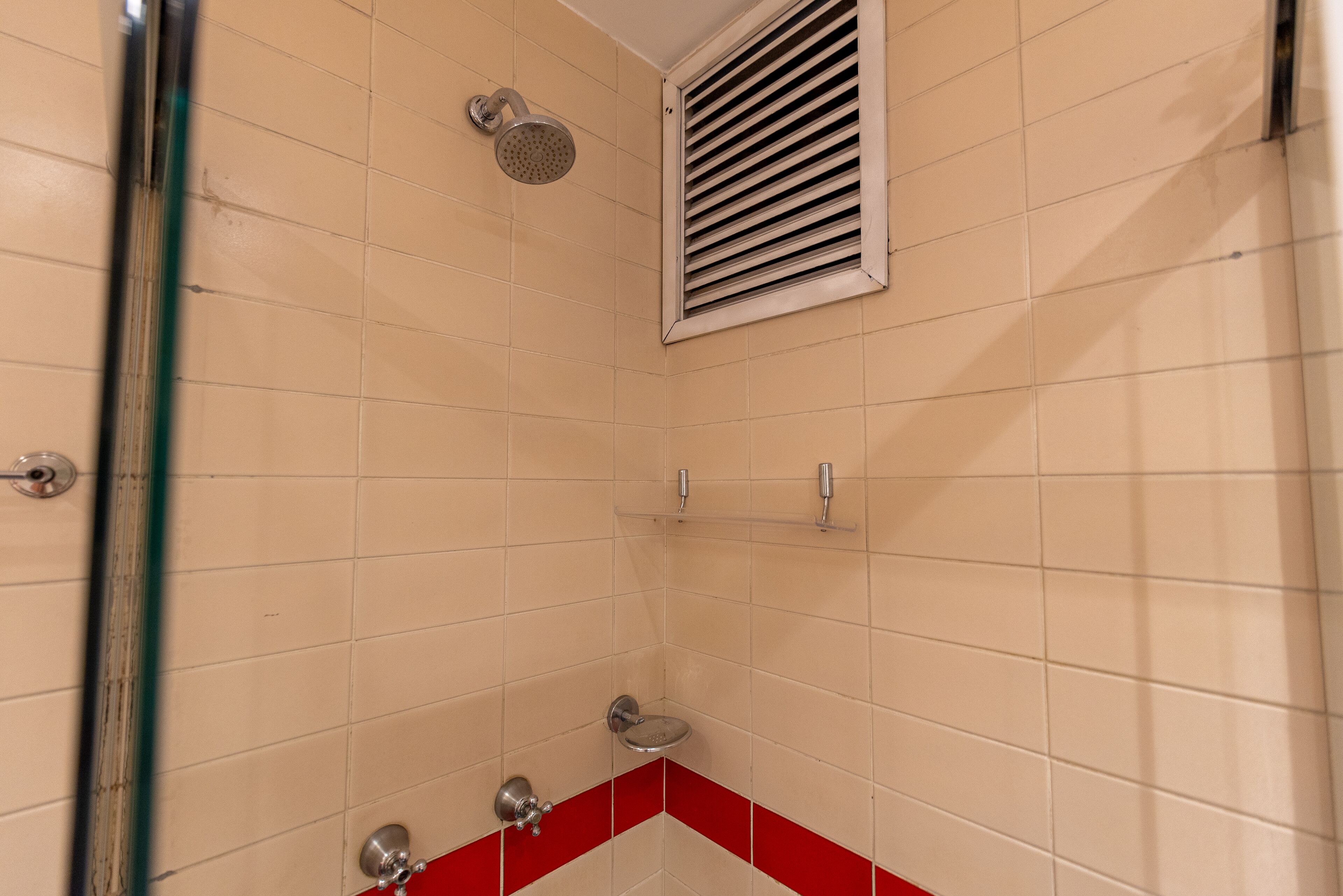 Casa de banho com azulejos bege, uma faixa vermelha, chuveiro com chuveiro manual, sanita e grelha de ventilação.