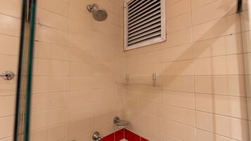 Casa de banho com azulejos bege, uma faixa vermelha, chuveiro com chuveiro manual, sanita e grelha de ventilação.