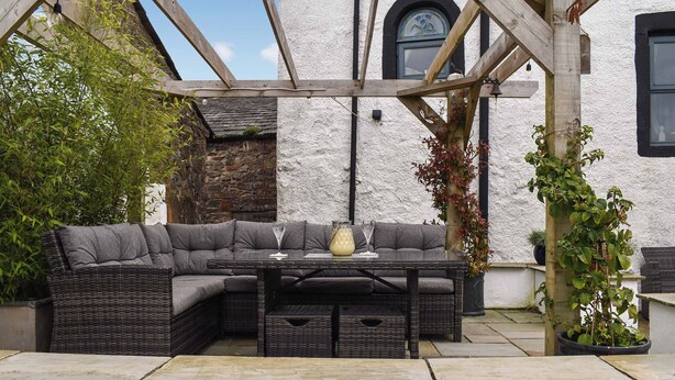 A covered outdoor seating area with a wicker sofa and a table, set against a white building with a stone wall and a window.