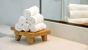 Ein Stapel weißer Handtücher auf einem hölzernen Hocker in einem Badezimmer mit Marmorplatte und einem Spiegel, der einen Handtuchhalter reflektiert.