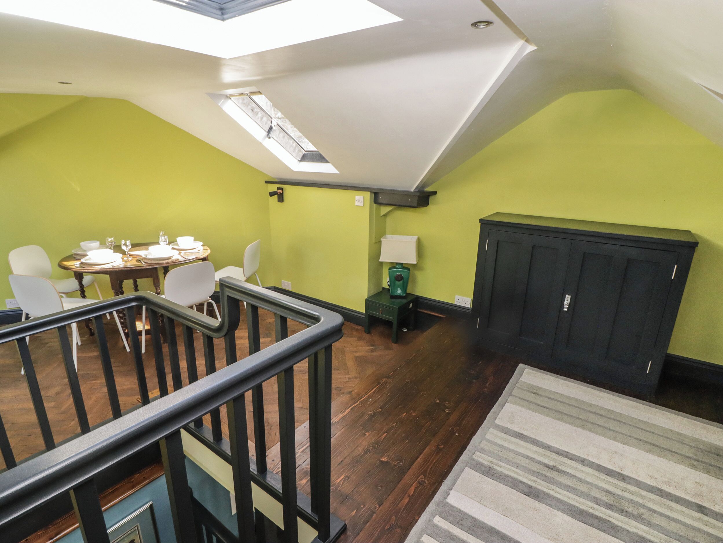 A dining area with a table set for four, a staircase, and a dark cabinet against a green wall.