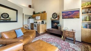 A living room with a leather sofa, a fireplace, a kitchen in the background, and a large painting on the wall. - Valley View (Flagstaff)