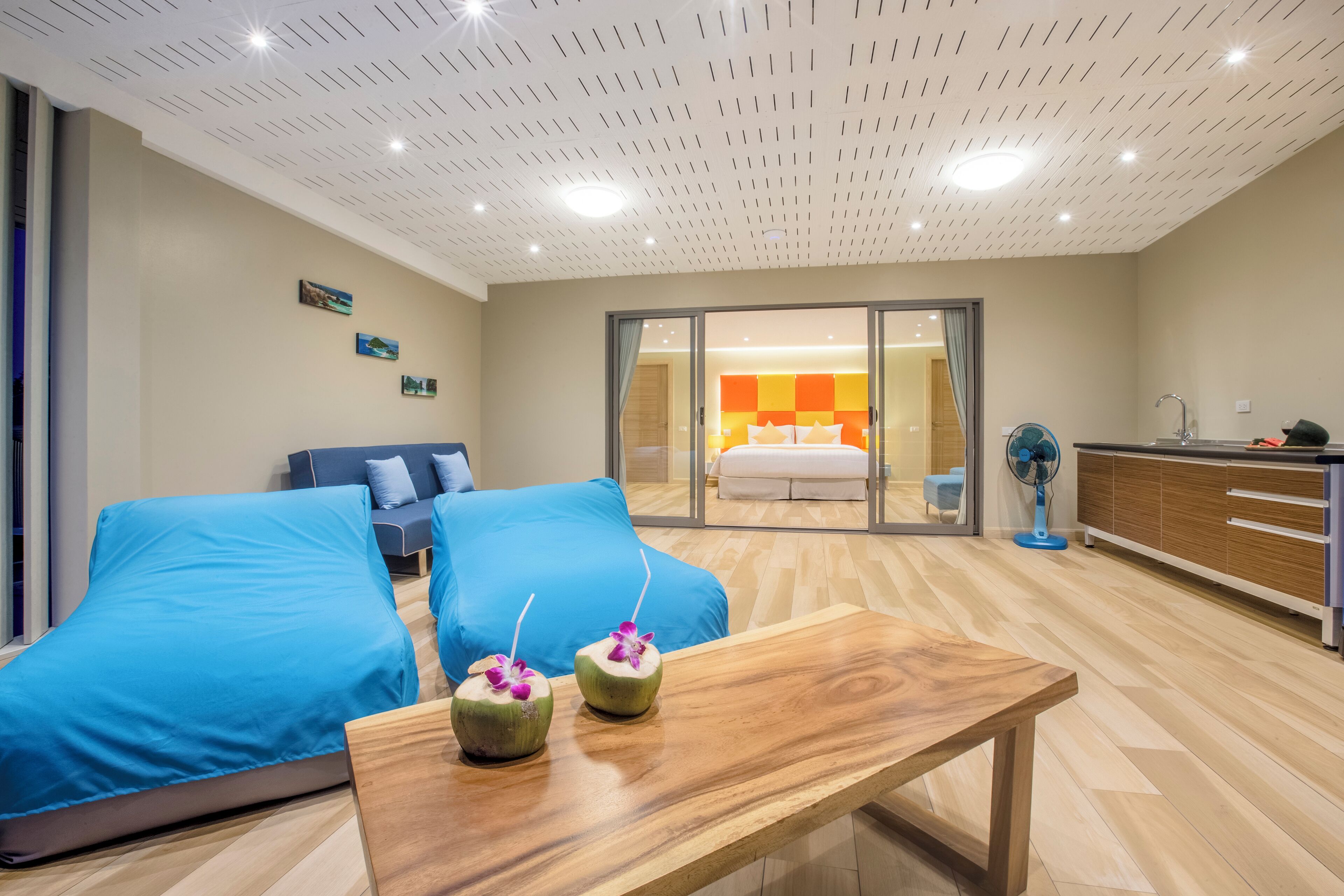 Habitación de hotel moderna con una cama grande, dos sofás azules, una mesa de madera y un ventanal corredero que da a un baño con una zona de ducha colorida.